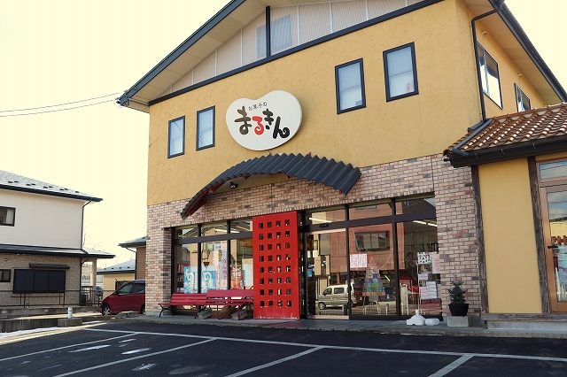 黄色い外壁にまるきんの看板、赤い入り口扉があり、店の前には駐車場がある店の外観写真