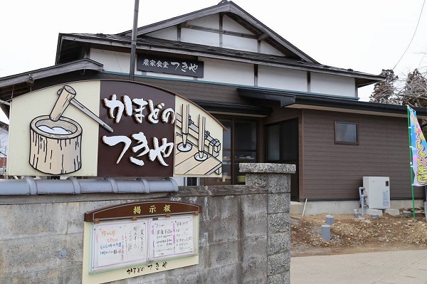 臼と杵で餅つきの柄が描かれている「かまどのつきや」の看板が門の塀に設置され、奥に平屋建て建物が見える和風造りの農家食堂 つきやの外観写真