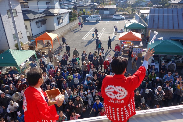 赤い法被を着た男性2名が建物の屋根から、下に集まっている来場者に餅まきをしている様子の写真