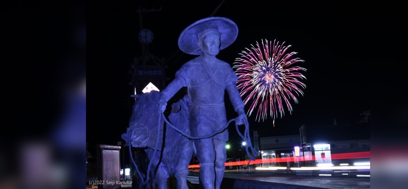 野田村フォトコンテストの小林フォト賞作品「よ市を輝す」撮影場所：道の駅のだ 牛方像前、背景に打ち上げ花火があり野田の牛方像が手前にある。
