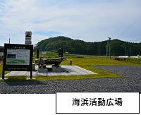 海浜活動広場：左側に案内板が立ち、手前の一部に砂利が敷かれ、奥には芝生が広がっている写真