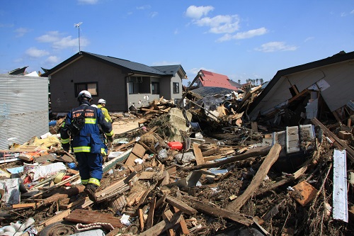 倒壊した家と、2階の高さまで積もった瓦礫の上を歩いている青い作業服と、黄色い作業服を着た人たちの後ろ姿を写した写真