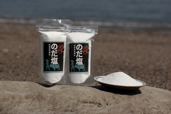 海辺にある岩の上に並べたパック入りの「のだ塩」が2個とお皿に盛られた「のだ塩」の写真