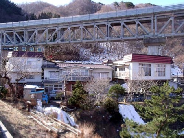 後方には山々と鉄道橋が通っており、屋根部分に「小野旅館」と書かれた看板が設置され、白い外壁で上部の一部が茶色に塗装されている小野旅館の外観写真