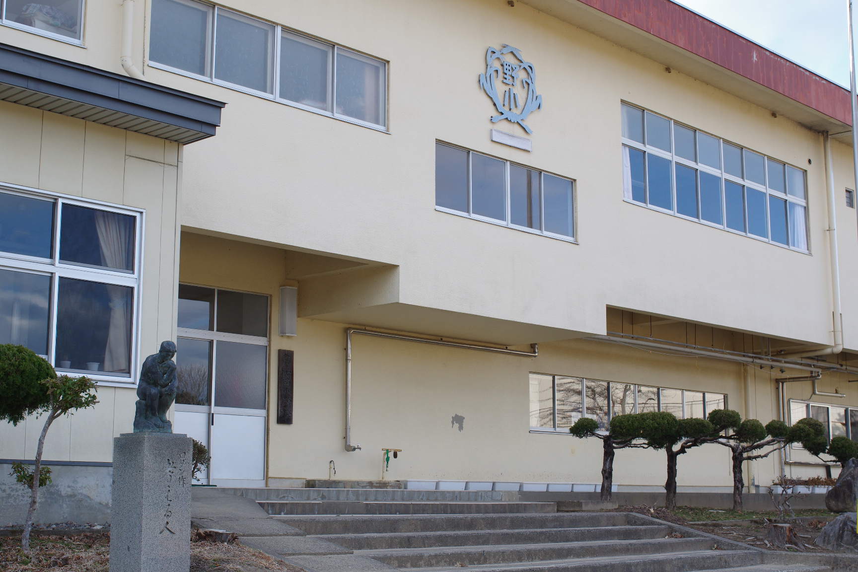 クリーム色の外壁で校章が表記されており、考える人の銅像が建物前に設置されている野田小学校の外観写真