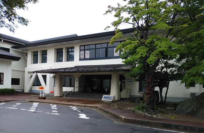 建物の入り口横に大きな木の植栽があり、二階建てで白い外壁の野田村総合センターの建物外観写真