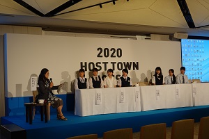 舞台に設置された席について話を聞いている野田中学校の生徒たちとその横でマイクで話をしている女性の写真