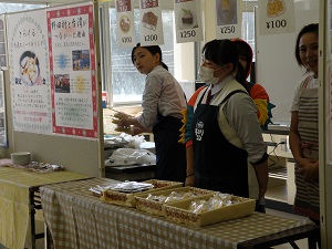 商品の写真と値段が書かれたメニューとテーブルに置かれた商品、販売している久慈工業高校料理部の生徒の写真