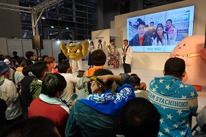 ステージ上の2体のキャラクターと野田村の代表者、スクリーンには映像が映し出され、その様子を見ている多くの来場者の写真