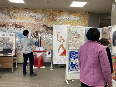 展示されている聖火リレートーチの右側に設置された掲示板に、縦二列に貼られたアートポスターの展示を見ている人達の写真