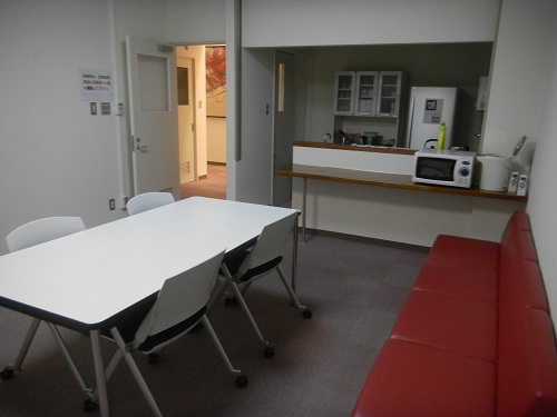 右の壁側に赤いソファー、中央に4人掛けのテーブルと椅子、 前方には冷蔵庫や食器棚、電子レンジや、電気ポットが設置されている娯楽談話室の写真