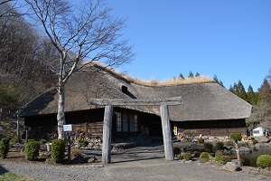 木製の鳥居のような門の奥に藁葺き屋根の古い平屋の建物がある南部曲り家の外観写真