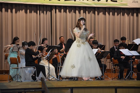 彰化市管弦楽団の前に立ち、白いドレスを着て歌を歌っている女性の写真
