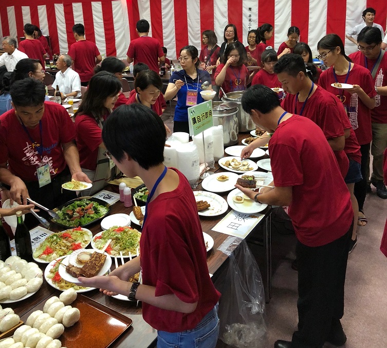 テーブルの上に並んだおにぎりや焼きそば、田楽、ホタテなどの料理をお皿にとっている彰化市管弦楽団員の写真