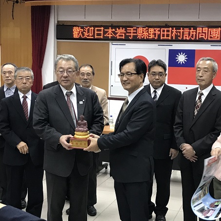野田村長と林市長が仏像の記念品を一緒に持っており、その周囲に関係者が立っている写真